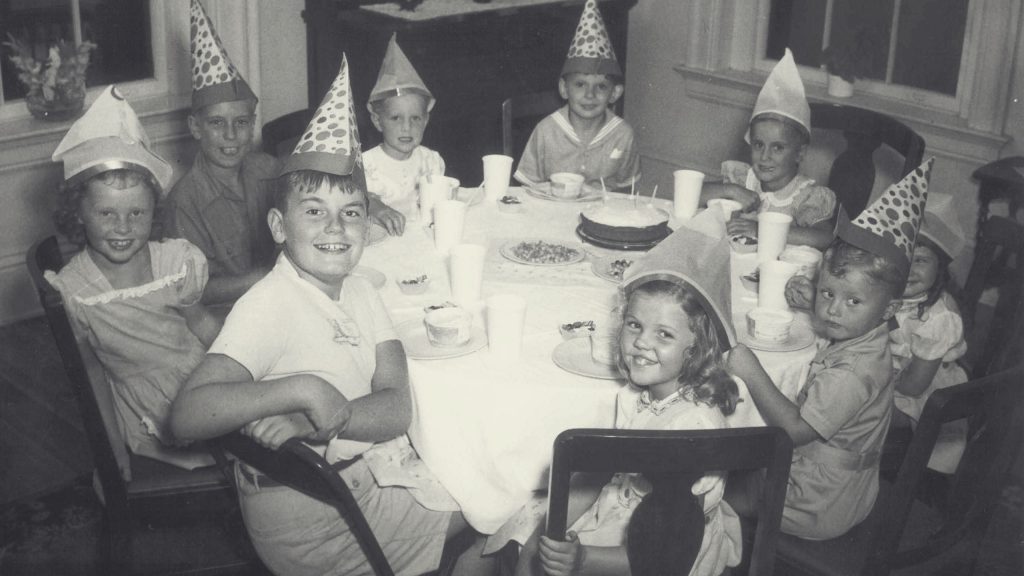 A black and white photo of nine children sitting around a table at a birthday party. They are wearing party hats and smiling. Plates, cups, and a cake are visible on the table. The children are dressed in casual clothing, and the room looks bright with windows in the background.