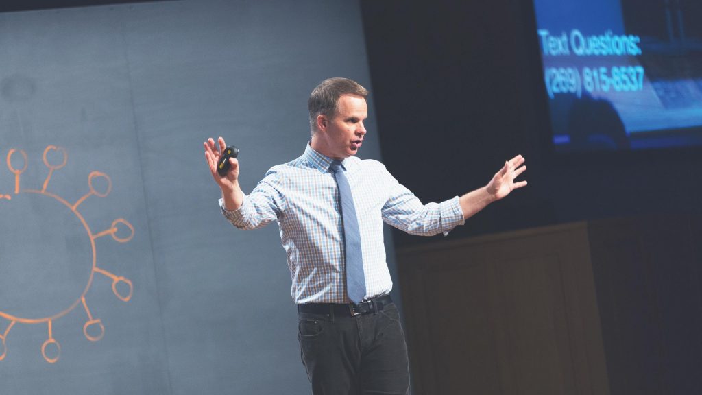 A man in a checkered shirt and tie is standing on a stage with a microphone headset, gesturing with both hands. Behind him is a screen displaying text and a graphic of a network diagram. The background is dimly lit.