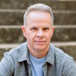 A middle-aged man with short, gray hair is looking directly at the camera with a slight smile. He is wearing a light blue shirt under a gray jacket. The background consists of blurred outdoor steps.