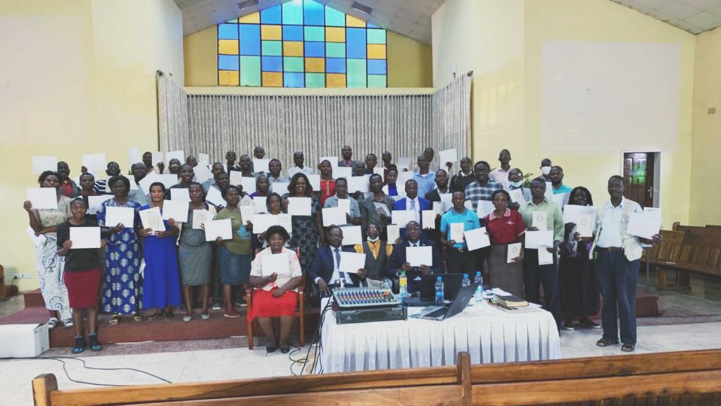 A large group of people poses for a photo inside a building, holding certificates. Behind them is a colorful stained glass window. In front, there is a table with electronic equipment and a few seated individuals. Some wooden benches are visible in the foreground.