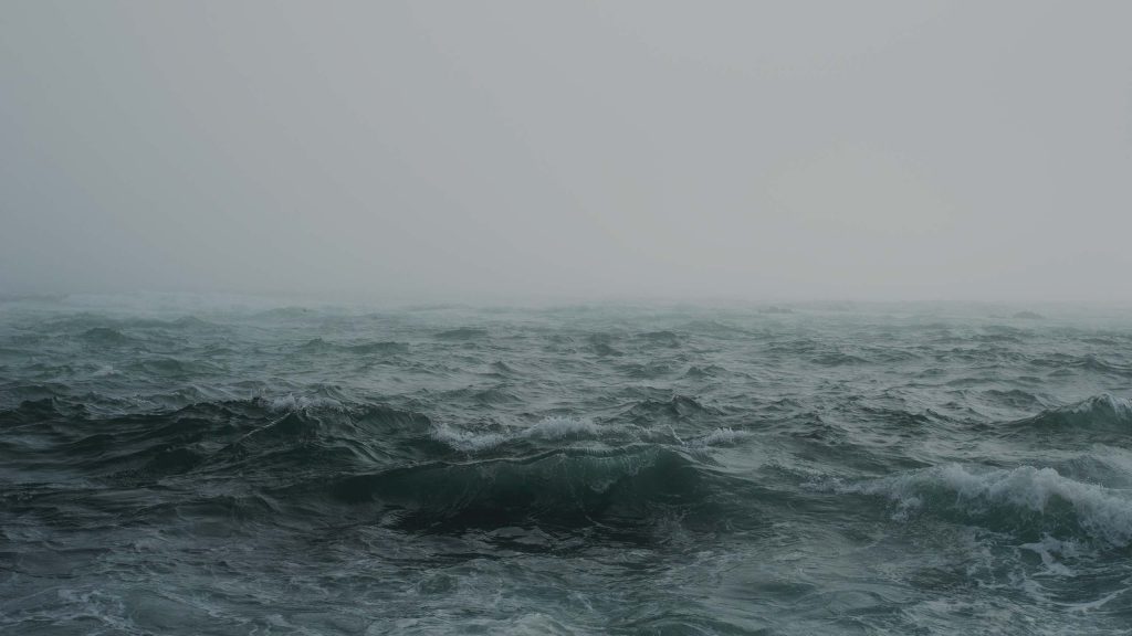A vast, tumultuous ocean is depicted with dark, choppy waves under a heavy, overcast sky, creating a moody, somber atmosphere. The horizon is barely visible due to the dense fog.